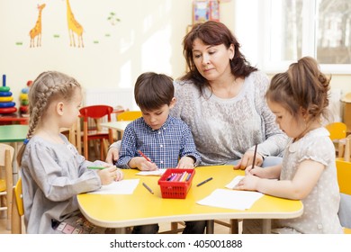 Three Preschooler, Two Girls And A Boy, Paint Pencils At The Table. Mature, Handsome Caucasian Brunette Woman, Caregiver, Watching And Smiling.