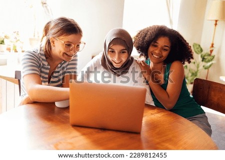 Three pretty female teenagers are looking at the laptop screen and laughing. Caucasian, afro american and muslim teen girls are watching a webinar or chatting at the computer