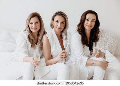 Three Pretty Women Sitting On A White Bed, Dressed All In White, Holding Champagne Glasses And Smiling