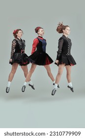 Three professional women of Irish dance ensemble in concert costumes and Ghillies Soft Shoes jump together in a row. Full-length studio portrait on a grey background. - Shutterstock ID 2310779909