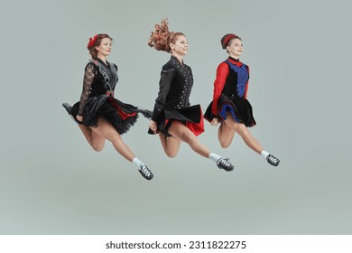 Three professional women of Irish dance ensemble in concert costumes and Ghillies Soft Shoes jump together in a row. Full-length studio portrait on a grey background. - Shutterstock ID 2311822275