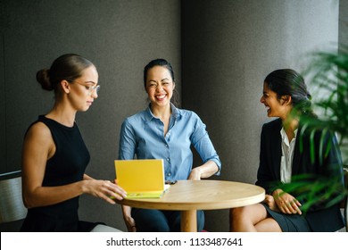 Three Professional Women Of Varying Ethnicities Sit And Have A Meeting Discussion Around A Table In A Meeting Room During The Day. One Is An Indian Asian Woman, The Other Chinese And The Last White.