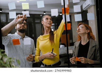 Three professionals engaged in a brainstorming session, using colorful sticky notes on a glass board to share ideas. They are focused and actively discussing their project.