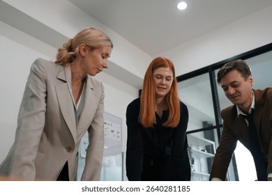 Three professionals gather around a table in a bright office, discussing plans and reviewing documents. They display focused expressions as ideas are exchanged.