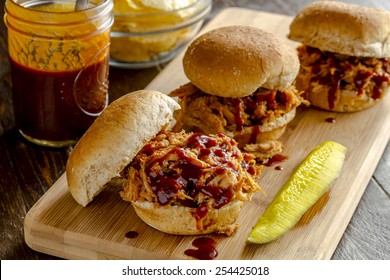 Three Pulled Pork Barbeque Sliders Sitting On Wooden Cutting Board With Dill Pickle With Bottle Of Sauce And Bowl Of Potato Chips