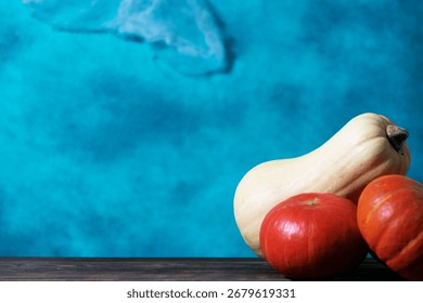 Three pumpkins, including two orange pumpkins and one pale yellow pumpkin, placed on a wooden surface against a bright blue textured wall with soft fabric details, an autumn harvest concept.