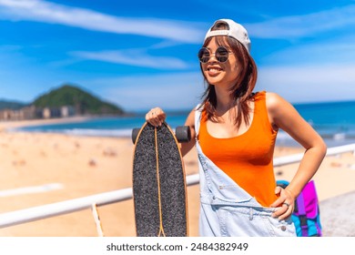 Three quarter length portrait of a chinese cool young woman with skateboard smiling standing next to the beach - Powered by Shutterstock