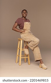 Three quarter length portrait of a Non-binary Afro-Latino in their 20s smiling with hands in pockets sitting on a stool on a neutral background. Stock Photo