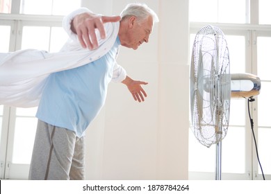 Three Quarter Side View Of A Senior Man With Arms Raised By The Fan Indoors Seeking Respite From The Heat.