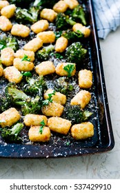 Three Quarters Shot Of Baked Tater Tots And Rich, Green Broccoli Topped With Grated Parmesan Cheese And Fresh Parsley In A Dark, Used Roasting Pan With Blue Striped Linen On Cream Marble Surface.