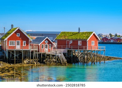 Three red cabins on stilts stand on a rocky outcropping in a Norwegian fjord, with turquoise water reflecting the clear blue sky. Reine, Lofoten, Norway - Powered by Shutterstock