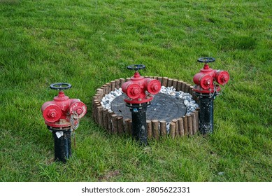 Three red fire hydrants stand on a green lawn beside a circular manhole cover surrounded by wooden edging and pebbles.