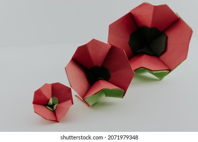 Three red origami poppies on a white background, two with black centers and green stem, one with green center, in a row from smallest to biggest