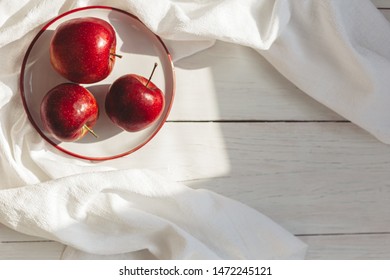 Three Ripe Red Apples On A White Plate. Top View On A White Wooden Table With Sunlight. Place For Text.