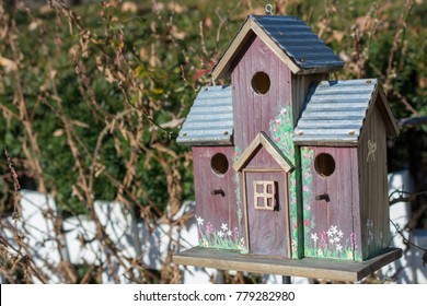 Three room two tier brown bird house sits along a white picket fence.