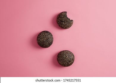 Three Round Crusty Charocal Black Burger Buns With Sesame Seeds On Rosy Background. Cuttlefish Ink Brioches For Making Sandwich, One Is Bitten. Fast Food And Nutrition Concept. View From Above