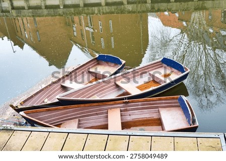 Three Rowing Boats Floating On A Calm River With Nearby Buildings Reflecting In The Water 