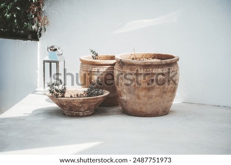 Three rustic clay pots with small plants are placed on a sunlit outdoor patio against a white wall. A metallic lantern sits beside, casting shadows and adding an elegant touch to a minimalist setting