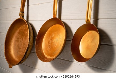 Three rustic copper fryingpans hanging on white kitchen wall in restaurant or at home. Round empty gold or yellow pans with handles. Kitchenware, utensils for cooking and frying, interior decoration
