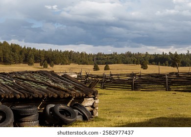 Three rustic log cabins sit in a mountain field among dense evergreens. The largest features a red door and green roof, while two smaller cabin, one partially collapsed, evoke rugged charm.