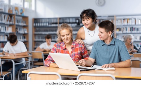 Three schoolchildren preparing for lessons in the school library are looking for information on a laptop and taking abstracts in a copybook - Powered by Shutterstock