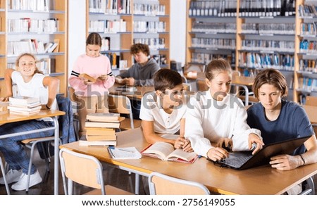 Three schoolchildren preparing for lessons in the school library are looking for the necessary information on a laptop, ..discussing topical issues