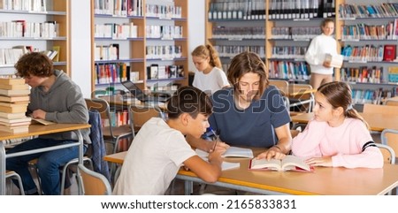 Three schoolchildren, who are engaged in lessons in the school library, make an synopsis of the necessary material from the ..textbook in an exercise book, discussing topical issues