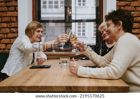 Three senior women friends having dinner at restaurant, attractive lady spending time together indoors. old friends drinking wine in modern apartment near window. Celebration concept.