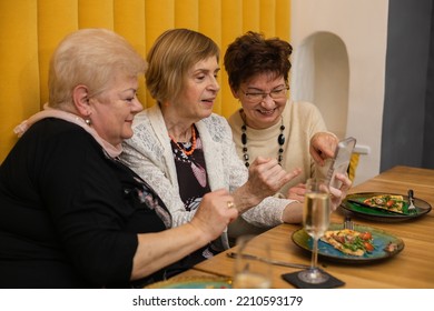 Three Senior Women Old Friends Relaxing And Having Dinner In Restaurant. Pensioners Sitting At Table In Cafe Room With Glasses Of Wine And Looking At Phone. Aged Attractive Ladies Celebrate Holiday.