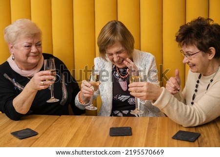 Three senior women old friends meeting together in restaurant. Pensioners sitting at table in room with yellow walls and joyfully drinking champagne. Aged attractive ladies celebrate holiday.