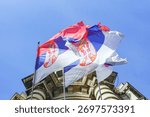 Three Serbian flags flutter in the wind on the façade of a building in the capital. National symbolism visualization and governmental legitimacy marker. Administrative authority representation.