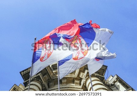 Three Serbian flags flutter in the wind on the façade of a building in the capital. National symbolism visualization and governmental legitimacy marker. Administrative authority representation.