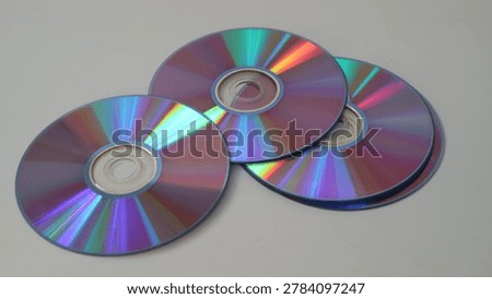 Three shiny compact discs with rainbow-colored reflections on a gray background. The discs are overlapping and have a metallic sheen.