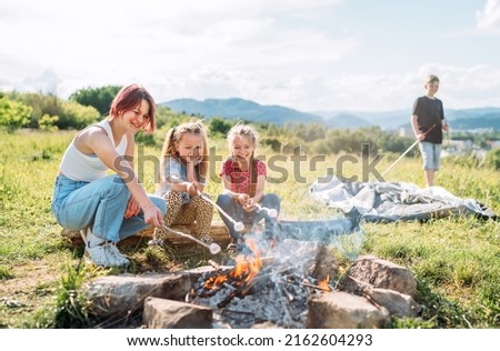 Three sisters cheerfully smiling while they roasting a  marshmallows candies on the sticks over the campfire flame. Happy family or outdoor picnic activities concept image.