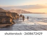 Three Sisters Rocks, New Zealand. Scenic view of a rugged coastline with tall sea stacks, gentle waves, and golden sunset light reflecting off the wet sand.