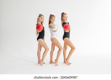 three slim artistic teenager girls in black and white leotards trains and having fun with balls on white background in rhythmic gymnastic exercise, children's professional sports