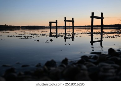 three small double jetties are reflected in the shallow water from which seaweed emerges and stones lie on the beach in the blurred foreground at sunset in the blue-orange horizon - Powered by Shutterstock