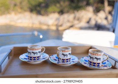 Three small patterned espresso cups filled with coffee sit on matching saucers arranged on a wooden serving tray on a boat. The porcelain cups feature
