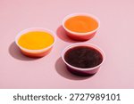 Three small plastic cups containing BBQ, mustard and spicy Roasted Red Pepper dipping sauce on pink background with some space for text or copy.