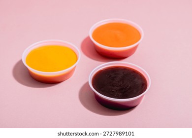Three small plastic cups containing BBQ, mustard and spicy Roasted Red Pepper dipping sauce on pink background with some space for text or copy.