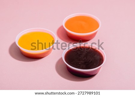 Three small plastic cups containing BBQ, mustard and spicy Roasted Red Pepper dipping sauce on pink background with some space for text or copy.