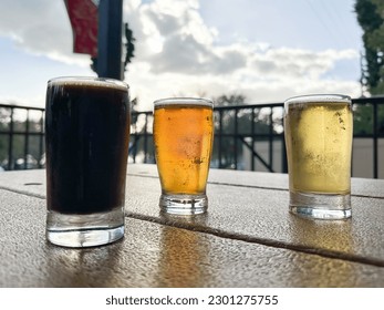 Three small sample glasses of cold craft beer. One glass is filled with a stout, the second is golden ale and the third is a lager. The brews are on patio table. The background is cloudy with blue sky