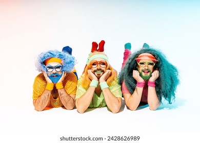 Three smiling drag queens in colorful 80s athletic outfits and wigs pose playfully on the floor against a light background.