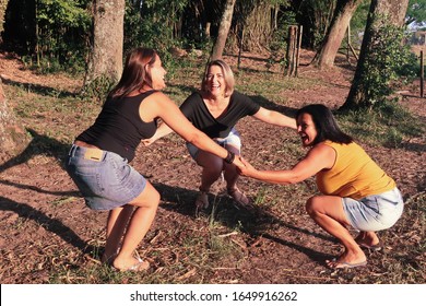 Three Smiling Friends, One With Blond Hair, One Brown And The Other Brunette, 30, 40 And 50 Years Old, Standing With Hands In Hand, Playing In A Circle, Happy