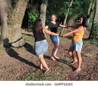 Three Smiling Friends, One With Blond Hair, One Brown And The Other Brunette, 30, 40 And 50 Years Old, Standing With Hands In Hand, Playing In A Circle, Happy