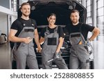 Three smiling workers, mechanics two men and woman in grey uniform standing at open hood, garage vehicle shop, auto mechanic technician maintenance customer car automobile at repair service.