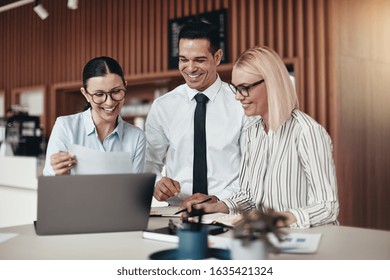 Three smiling young businesspeople standing together at a table in an office working on a laptop and going over paperwork - Shutterstock ID 1635421324