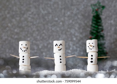 Three snowmen made from marshmallows and placed in silvery bokeh. A tree faded into the background and big smiles make them ready for the winter holidays. Horizontal, copy space
