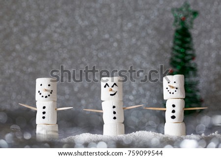 Three snowmen made from marshmallows and placed in silvery bokeh. A tree faded into the background and big smiles make them ready for the winter holidays. Horizontal, copy space