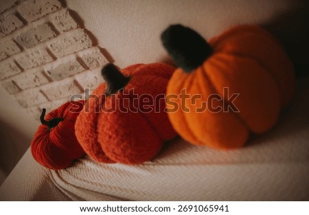 Three soft knitted pumpkins in warm orange tones placed on a cozy fabric surface near a textured wall, symbolizing autumn home decor and seasonal comfort.